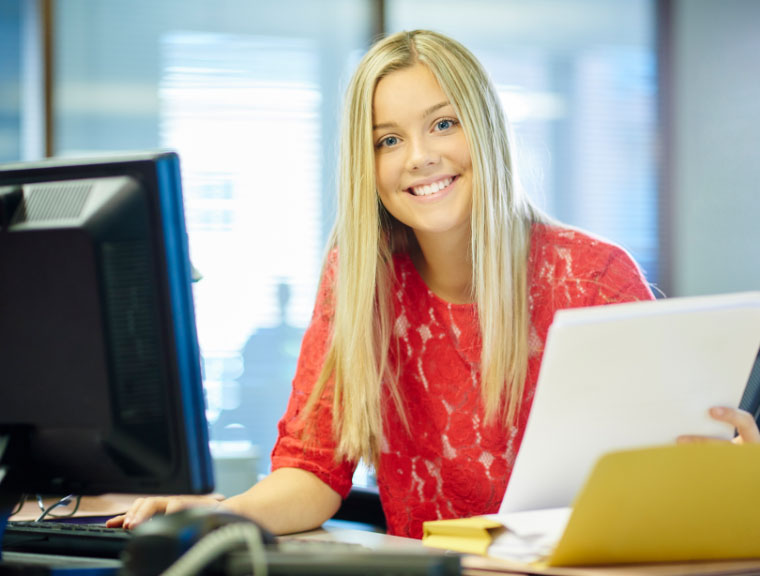 Smiling intern sitting at computer holding papers